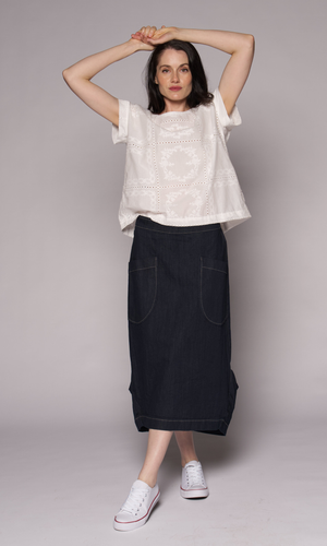 A woman modelling a white short-sleeve embroidered top and a dark navy denim midi skirt with front patch pockets, paired with white sneakers.
