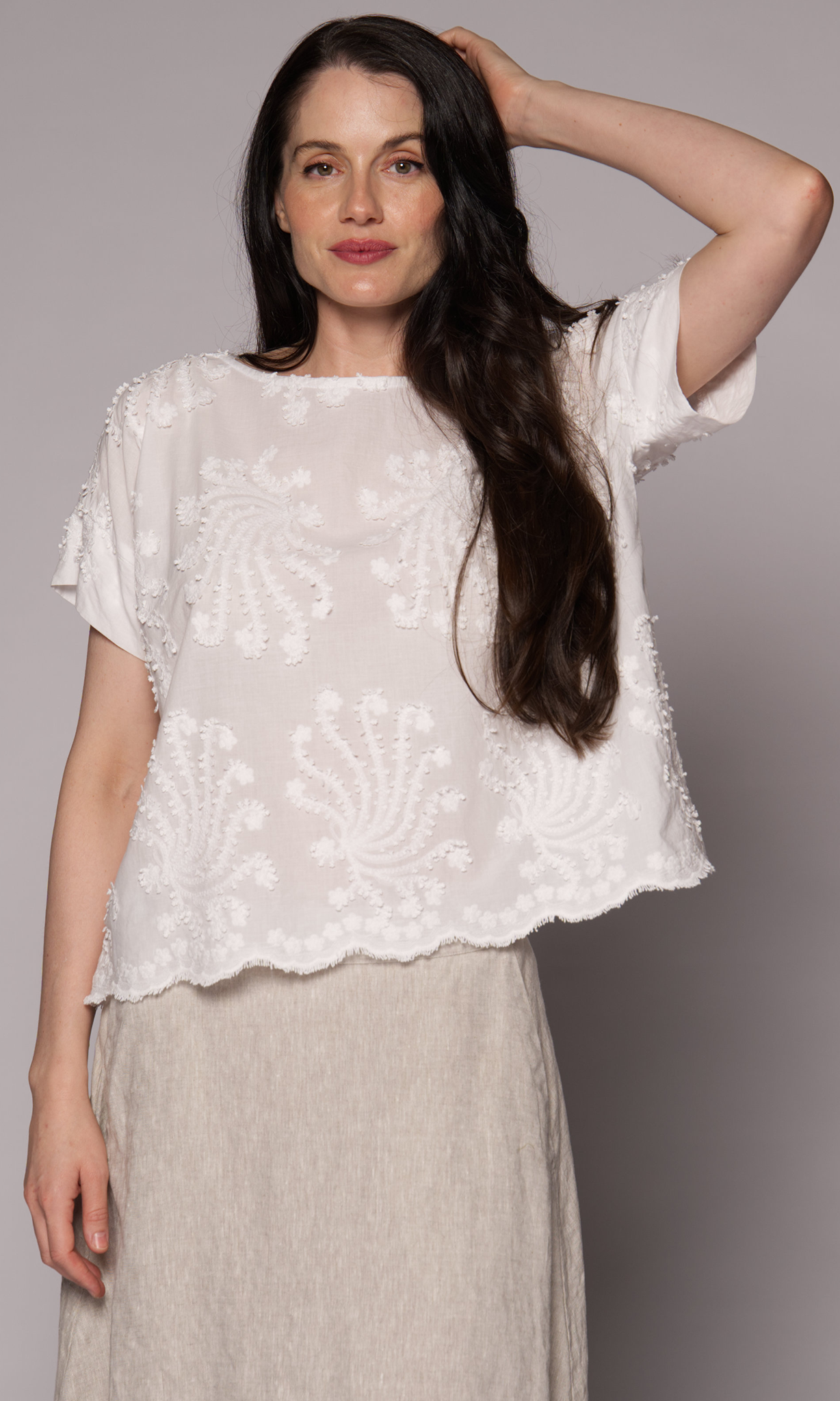 Dark brown haired woman modelling a white patterned top with one hand touching her head.