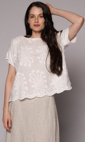 Dark brown haired woman modelling a white patterned top with one hand touching her head.