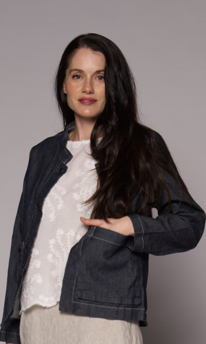 A woman with long dark hair wearing a dark grey denim jacket over a white lace top and a light beige skirt.