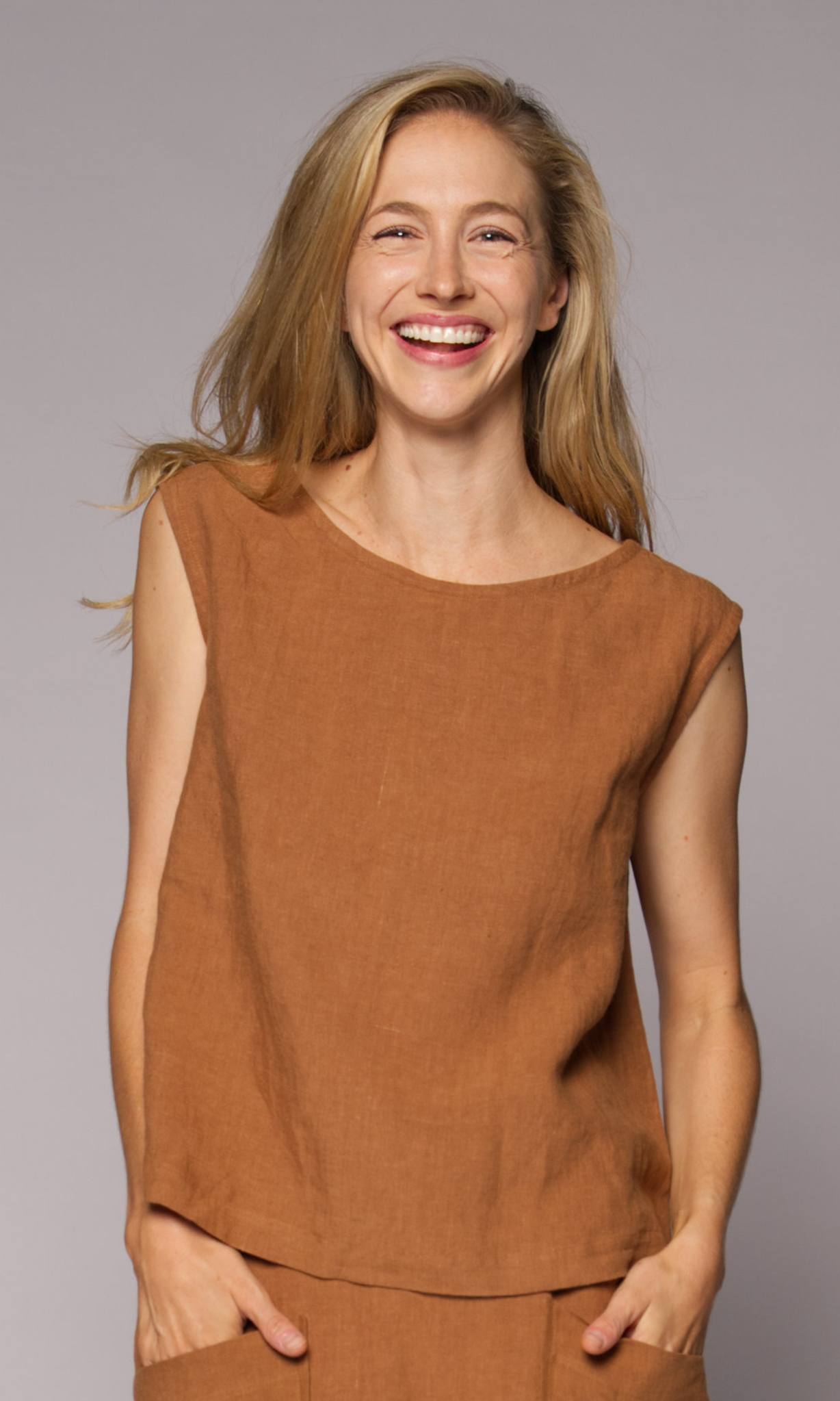 A smiling blonde woman wearing a sleeveless rust-brown linen top and matching skirt with hands in pockets.