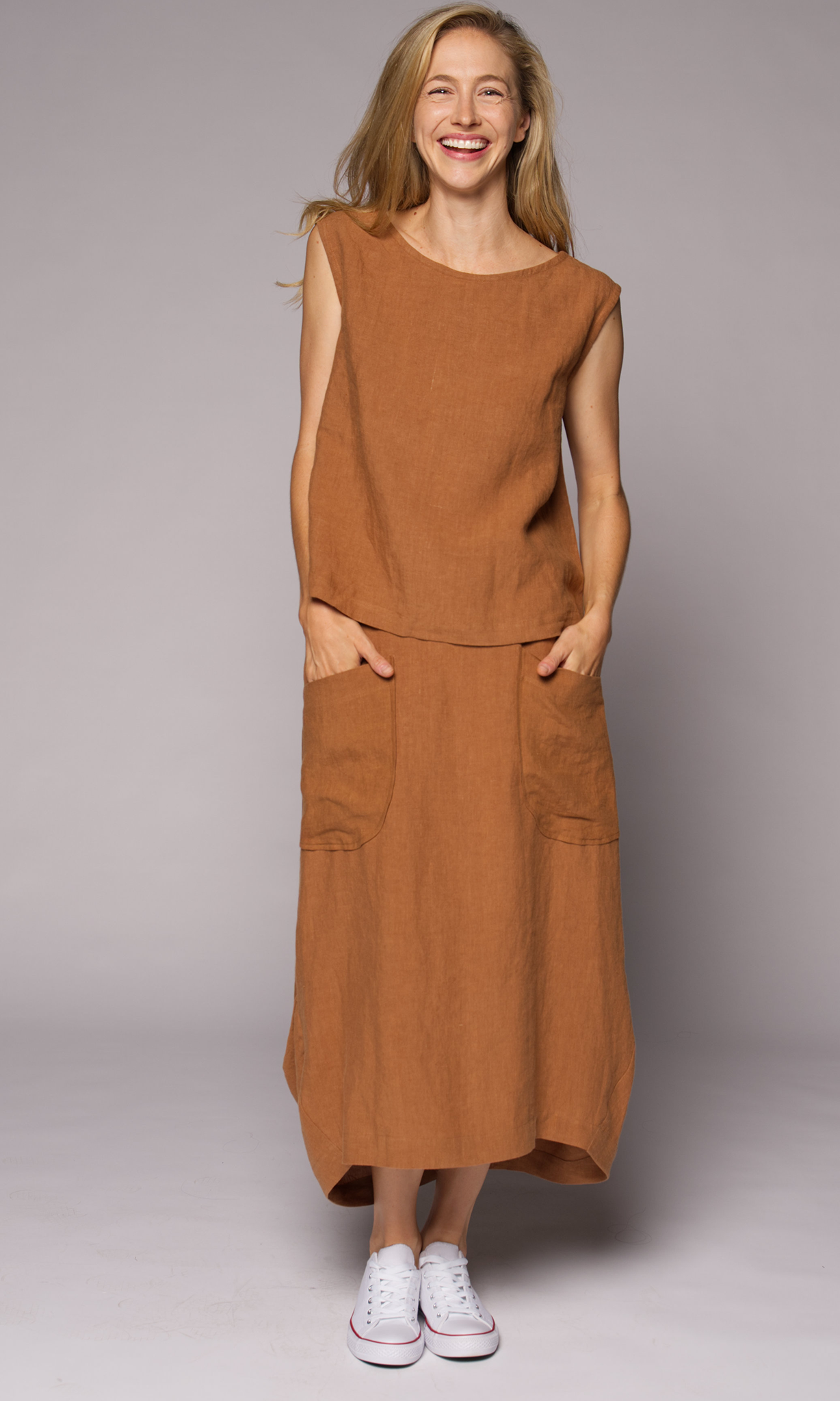 A woman modelling a sleeveless, rust-brown linen top and matching midi skirt with front patch pockets, paired with white sneakers.