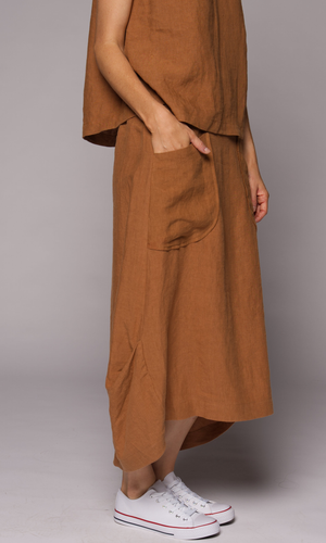 A person wears a loose, earthy-brown linen top and matching skirt with a large front patch pocket, paired with white low-top sneakers.