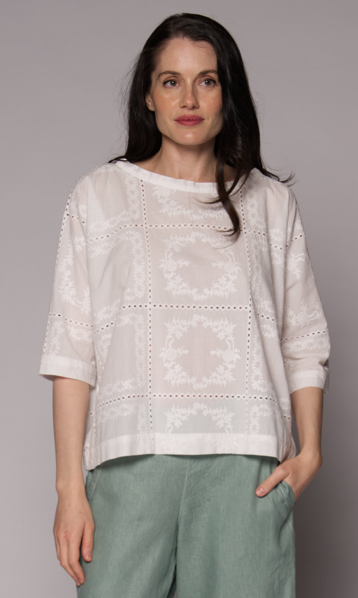 Dark haired woman with a 3/4 sleeve patterned top in white with light green pants.