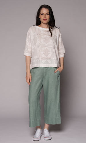 Dark haired woman with a 3/4 sleeve patterned top in white with light green pants.