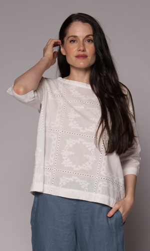 Dark haired woman with a 3/4 sleeve patterned top in white with light blue pants.