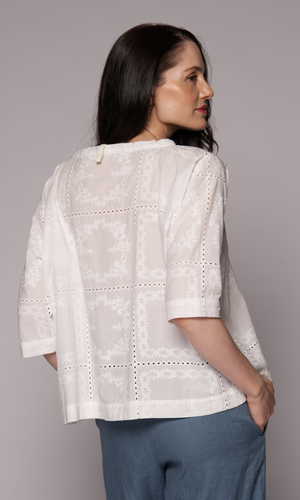 Dark haired woman with a 3/4 sleeve patterned top in white with light blue pants, turned to show us her back.