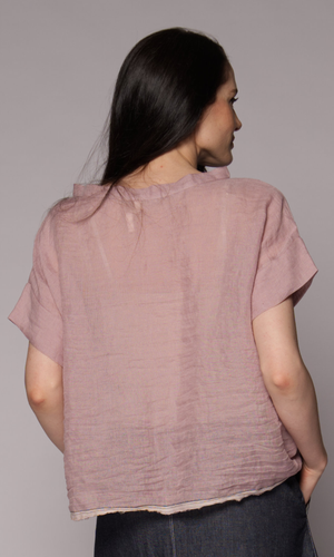 Dark brown haired woman modelling a light pink linen top and denim skirt with hand in pocket, showing the back of the top.