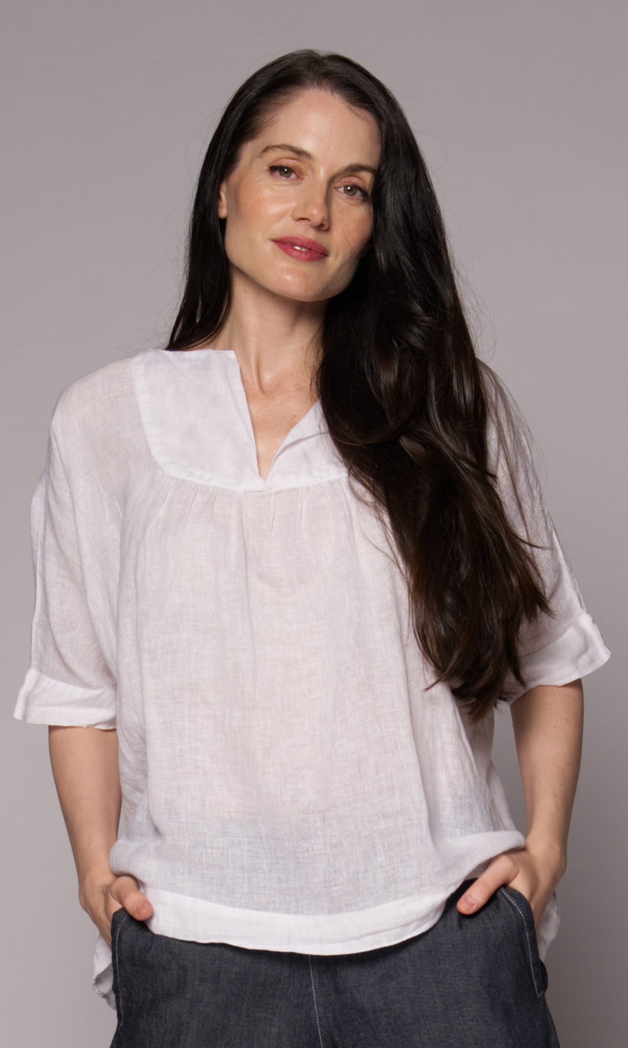 Brown-haired woman modelling a white linen top and denim shorts, with hands in her pockets.