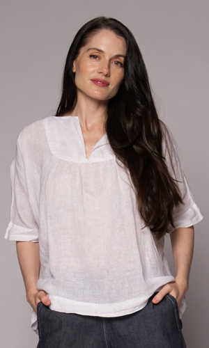 Brown-haired woman modelling a white linen top and denim shorts, with hands in her pockets.