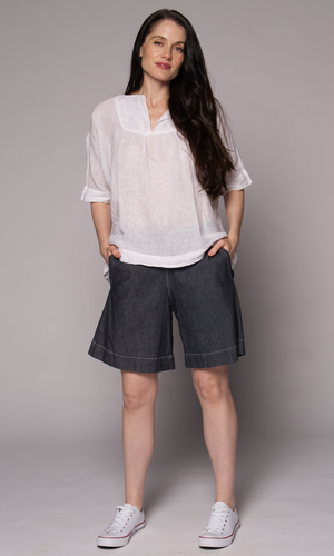 Brown-haired woman modelling a white linen top and denim shorts, with hands in her pockets.