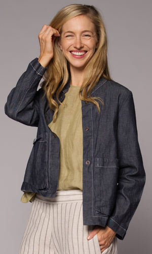 A woman with long blonde hair wearing a dark grey denim jacket over a yellow top and a light beige pin-striped pants.
