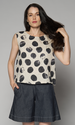 A woman models a sleeveless top with a black-and-white abstract polka-dot pattern, paired with dark denim shorts.