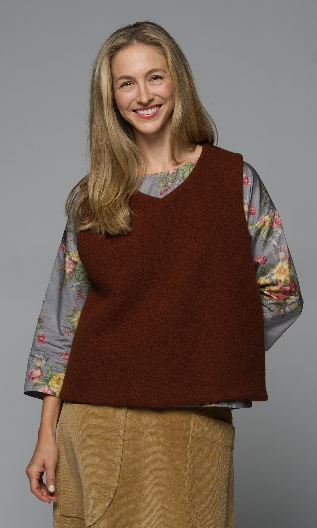 A woman in a rust coloured wool vest with a floral top and a caramel coloured skirt.