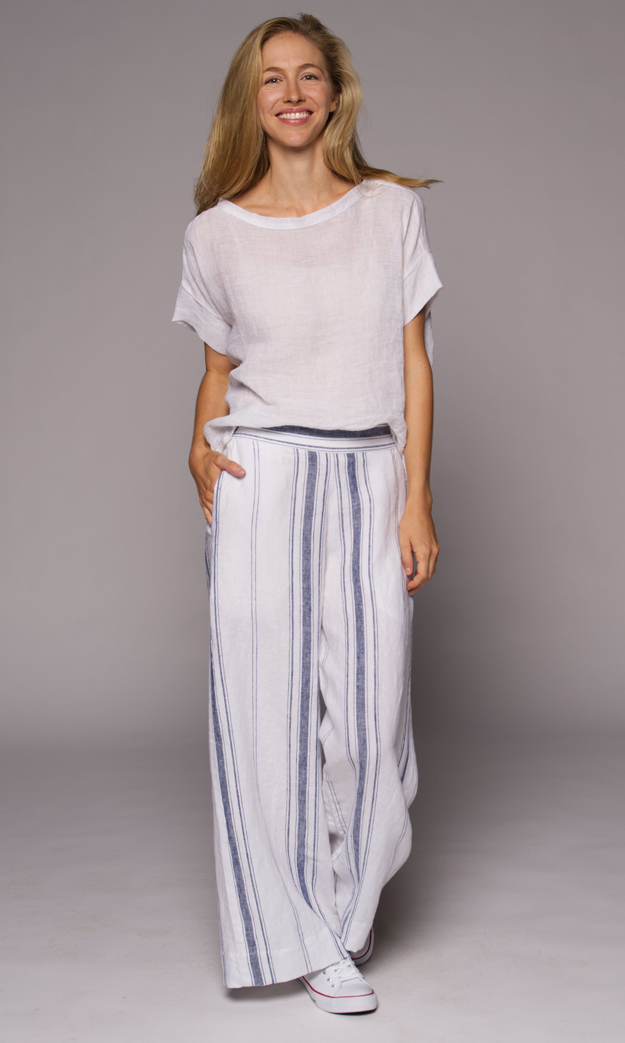 Blonde woman modelling a white linen top with blue striped linen pants, hand in pocket.