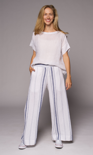 Blonde woman modelling a white linen top with blue striped linen pants, hand in pocket, standing with legs wide apart.