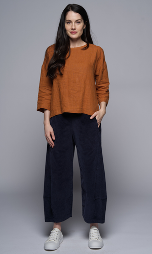 A woman wears a long sleeve rust coloured top and dark blue pants, with her hand in the pocket.