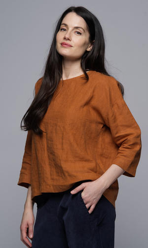 A woman wears a long sleeve rust coloured top and dark blue pants, with her hand in the pocket.