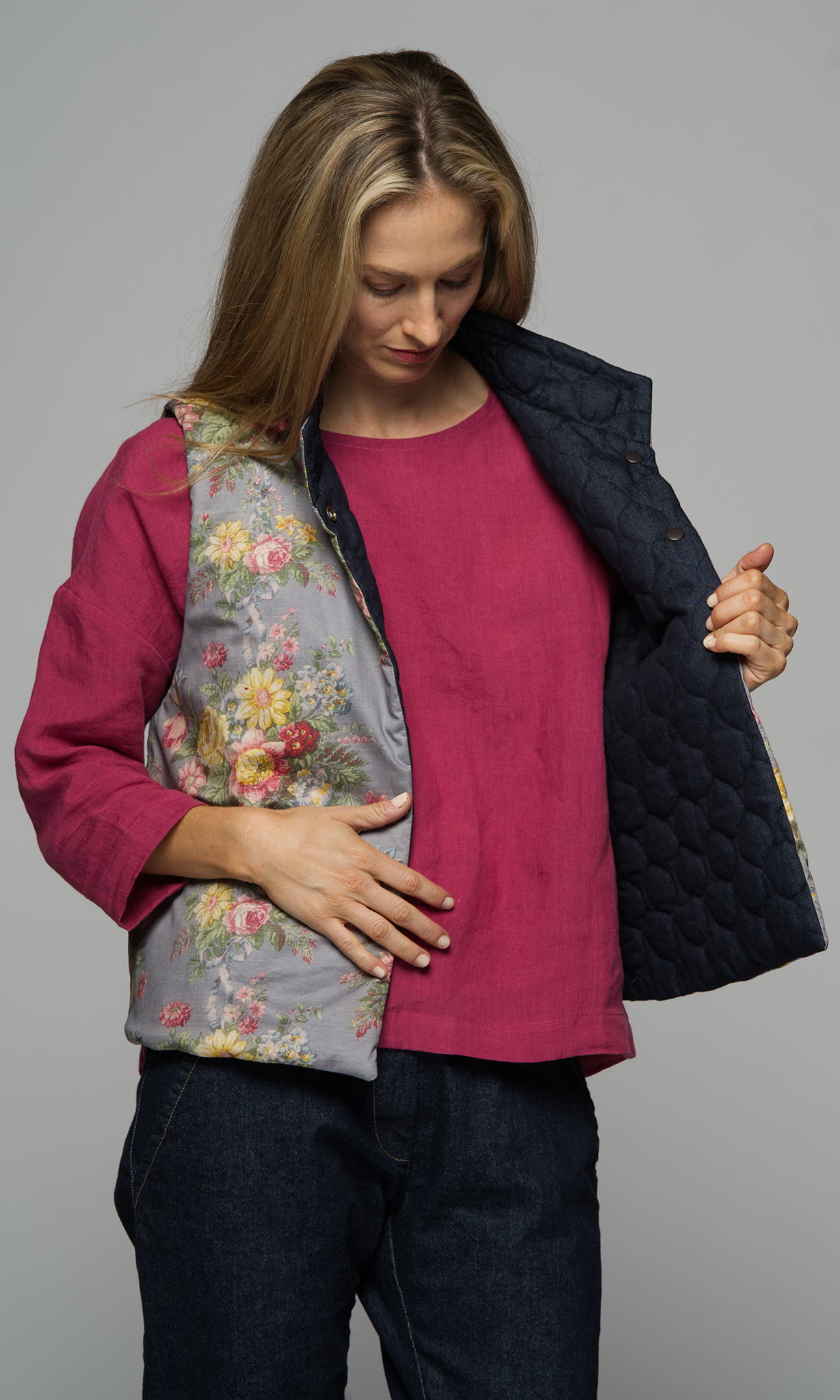 A woman wearing a floral vest with a pink top and a pair of blue jeans.