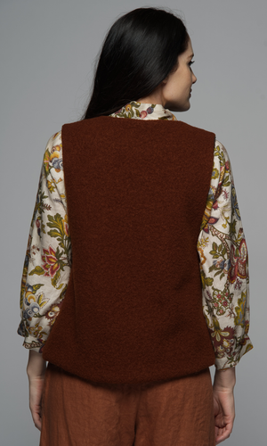 A woman in a rust coloured wool vest with a floral top and brown pants.