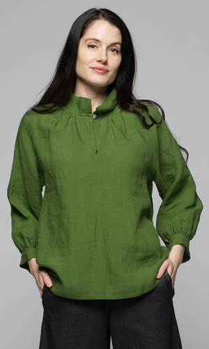 A woman wearing a long sleeve green top with black pants, with hands in pockets.