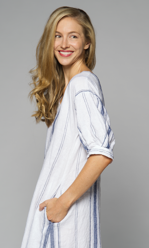 Blonde haired woman modelling a long striped linen dress turned to her side, close up of her hand in side pocket.