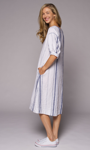 Blonde haired woman modelling a long striped linen dress turned to her side, hand in side pocket.