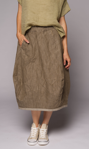 A person wears a loose, high-waisted brown linen skirt with side pockets, paired with a matching olive green top and cream sneakers, with one hand in a pocket.