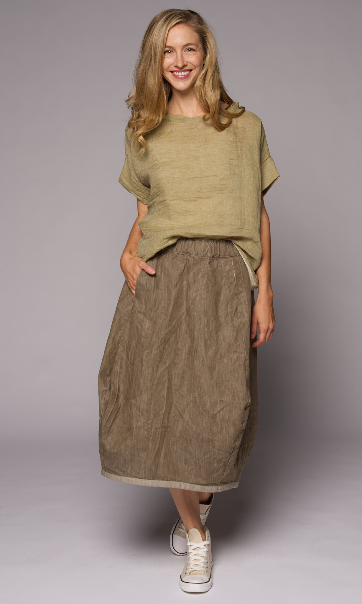 Blonde woman modelling a green linen top with a brown loose skirt, hand in pocket.