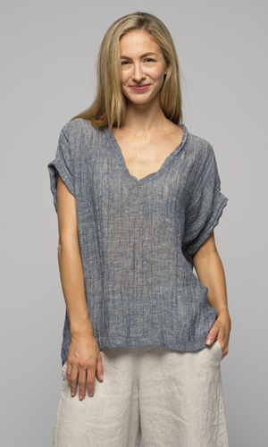 Woman wearing a blue/grey top with beige/ white linen flowing pants, facing the camera, smiling.