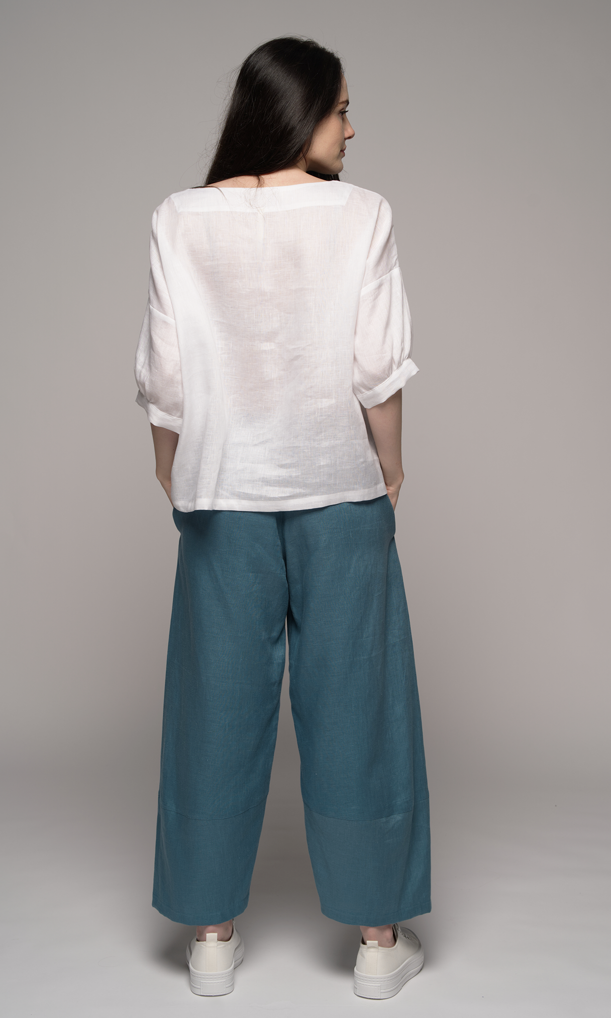 Lady wearing a white top with sleeves to elbows and a teal blue coloured ankle length pants, showing back of clothing.