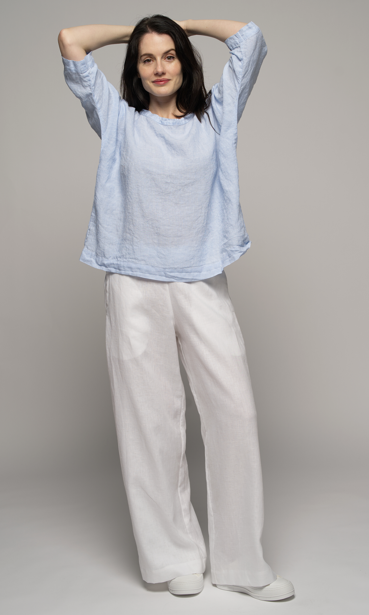 Woman wearing a light blue top and white pants, arms crossed above her head.