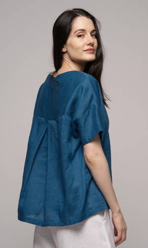 Woman wearing a blue blouse with white pants, facing to the right, showing you back of blouse.