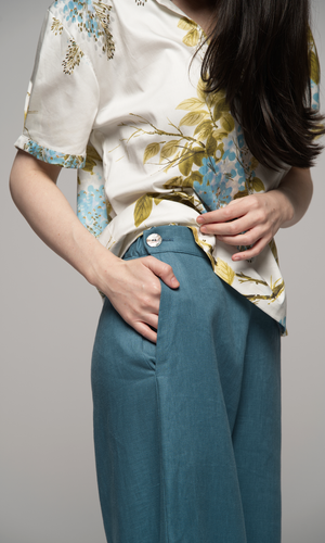 Woman wearing a floral blouse and teal pants showing closeup of pocket and button detail.
